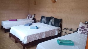 Une chambre avec trois lits simples, chacun agrémenté d’un oreiller à motifs différents. On y trouve un petit banc en bois et une étagère fixée au mur, sur laquelle est posé un panier.