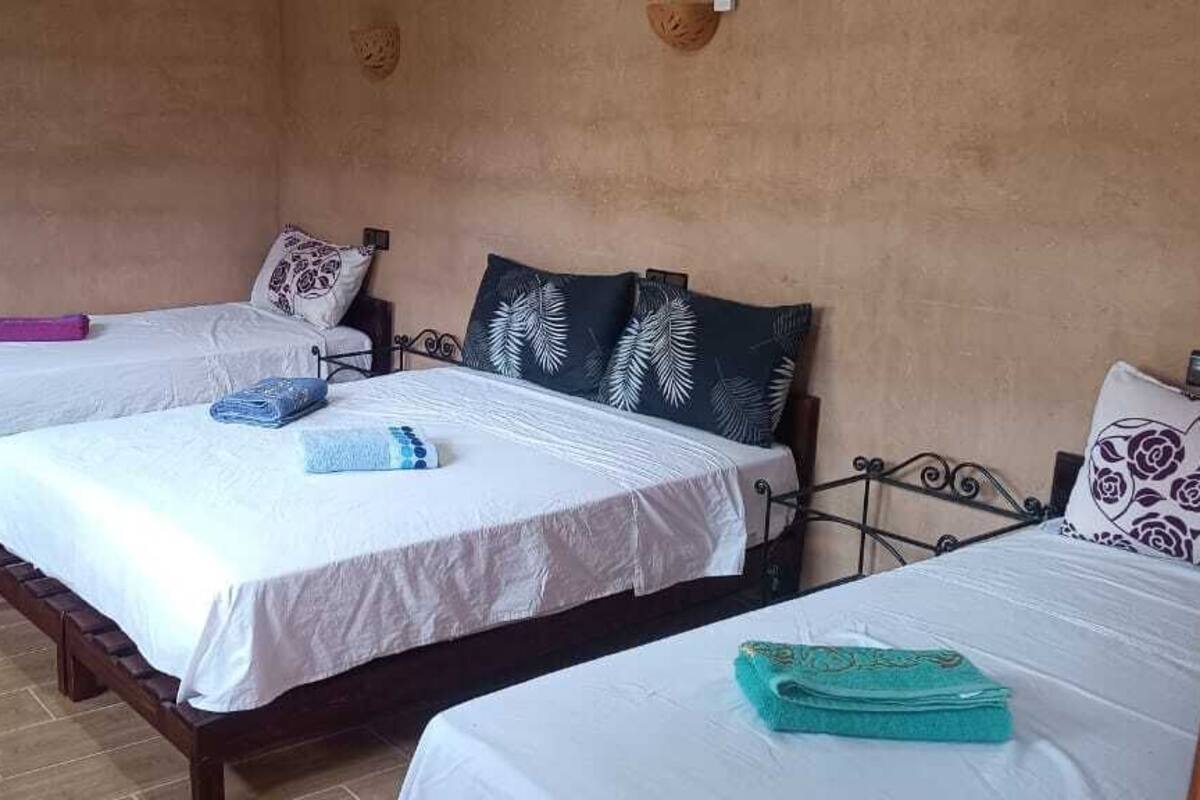 Ein Zimmer mit drei Einzelbetten, jedes mit einem verschiedenen gemusterten Kopfkissen. Es gibt eine kleine Holzbank und ein an der Wand befestigtes Regal mit einem Korb.