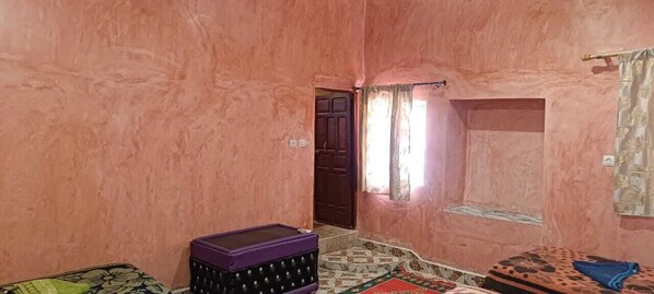 A room with pink walls, a wooden door, a window with curtains, a purple storage box, and a patterned rug. - Tissili Auberge Restaurant Oasis de Fint (Tarmigt)