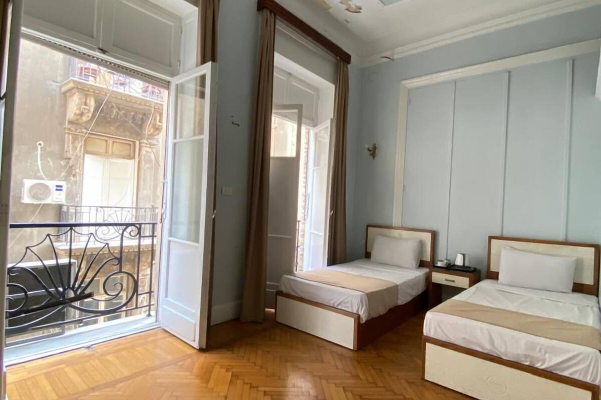 Habitación de hotel con dos camas individuales, suelo de madera con diseño espiga, balcón con barandilla de hierro forjado y ventilador de techo.