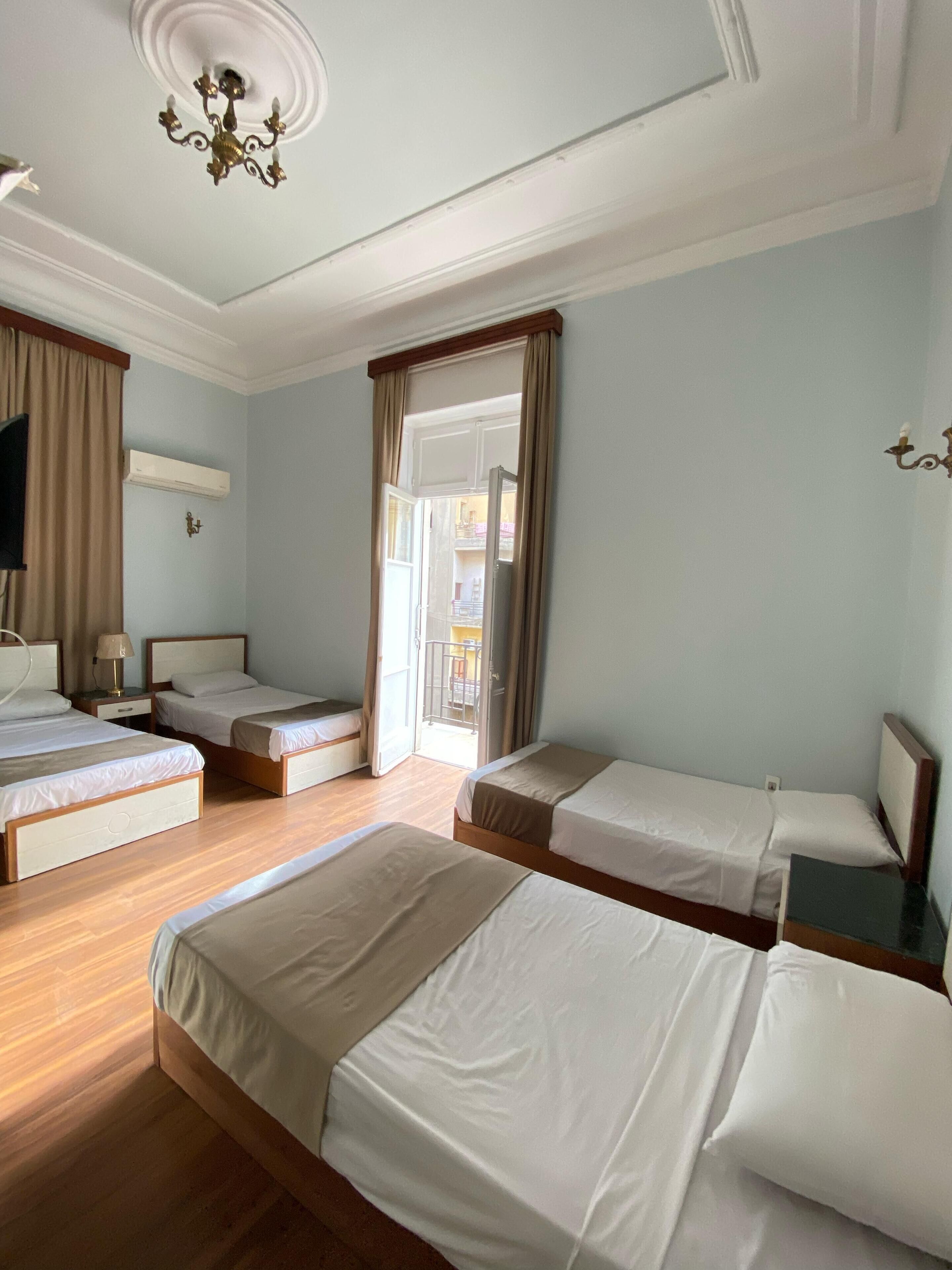 Una habitación de hotel con cuatro camas individuales, suelo de madera, un candelabro en el techo y un aire acondicionado, además de vistas al balcón exterior.
