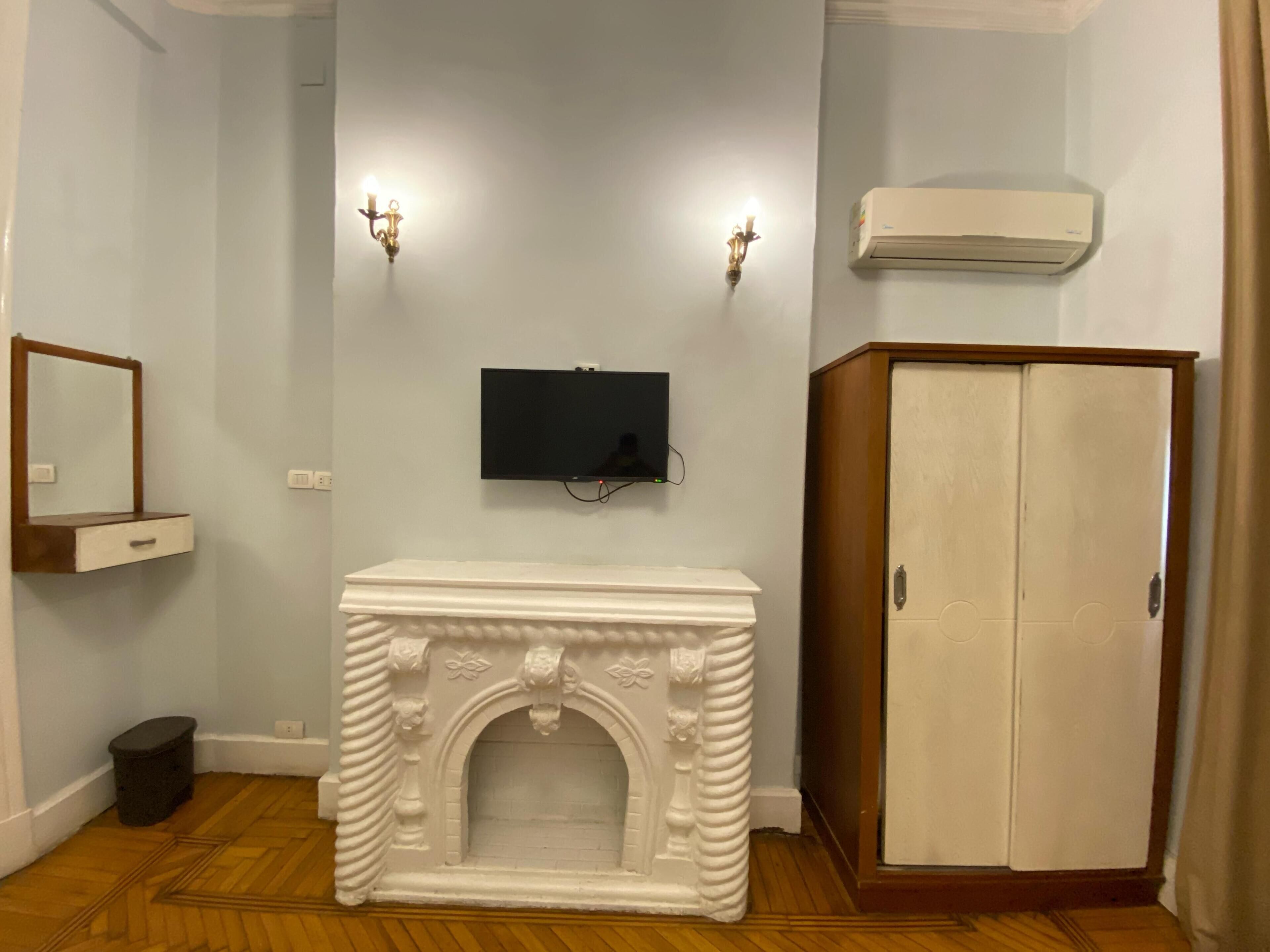 Una habitación con chimenea blanca, televisor montado, dos apliques de pared, un armario y un cubo de basura pequeño.