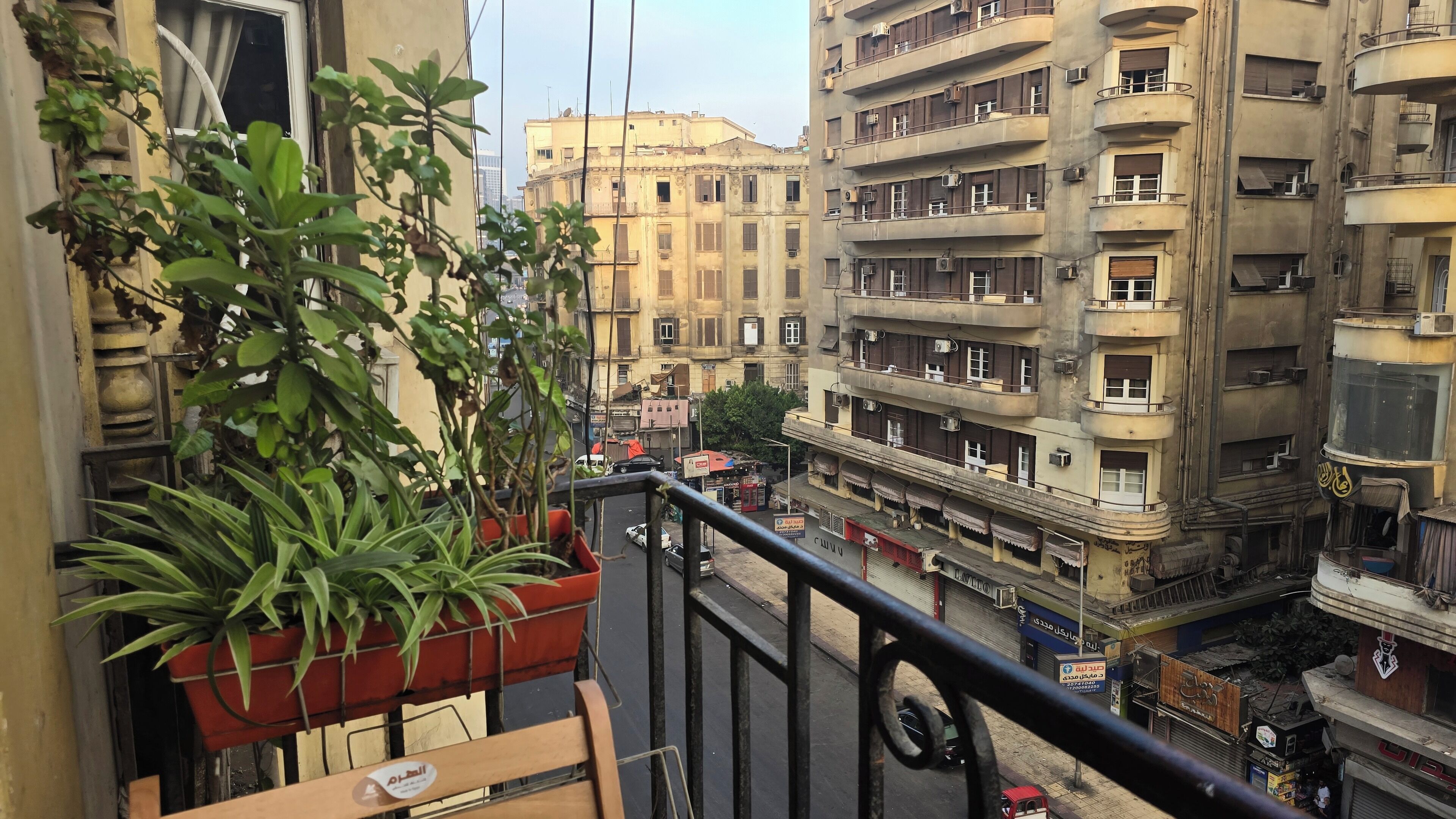 Un balcón con plantas en macetas, vista de una calle concurrida y edificios, y cielo despejado.
