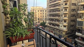 Een balkon met bloempotten, uitzicht op een drukke straat met gebouwen, en een heldere lucht.