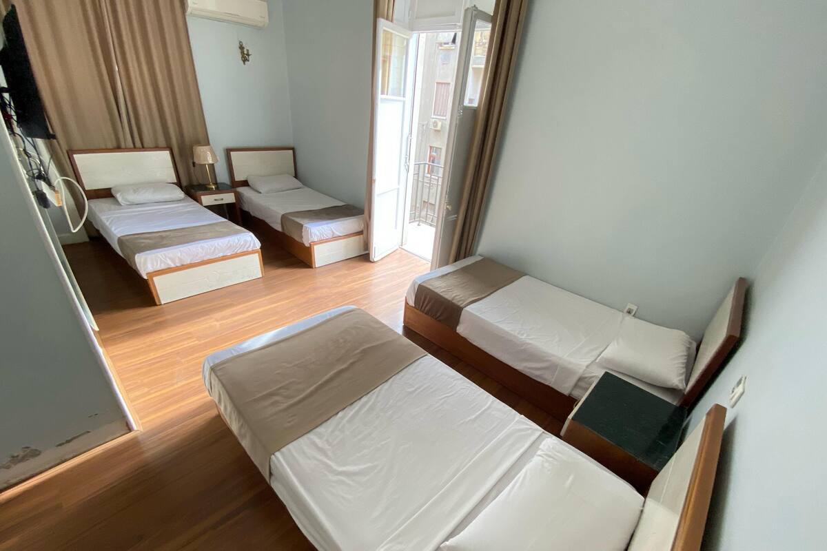 Habitación de hotel con tres camas individuales, piso de madera, ventana con cortinas y aire acondicionado montado en la pared.
