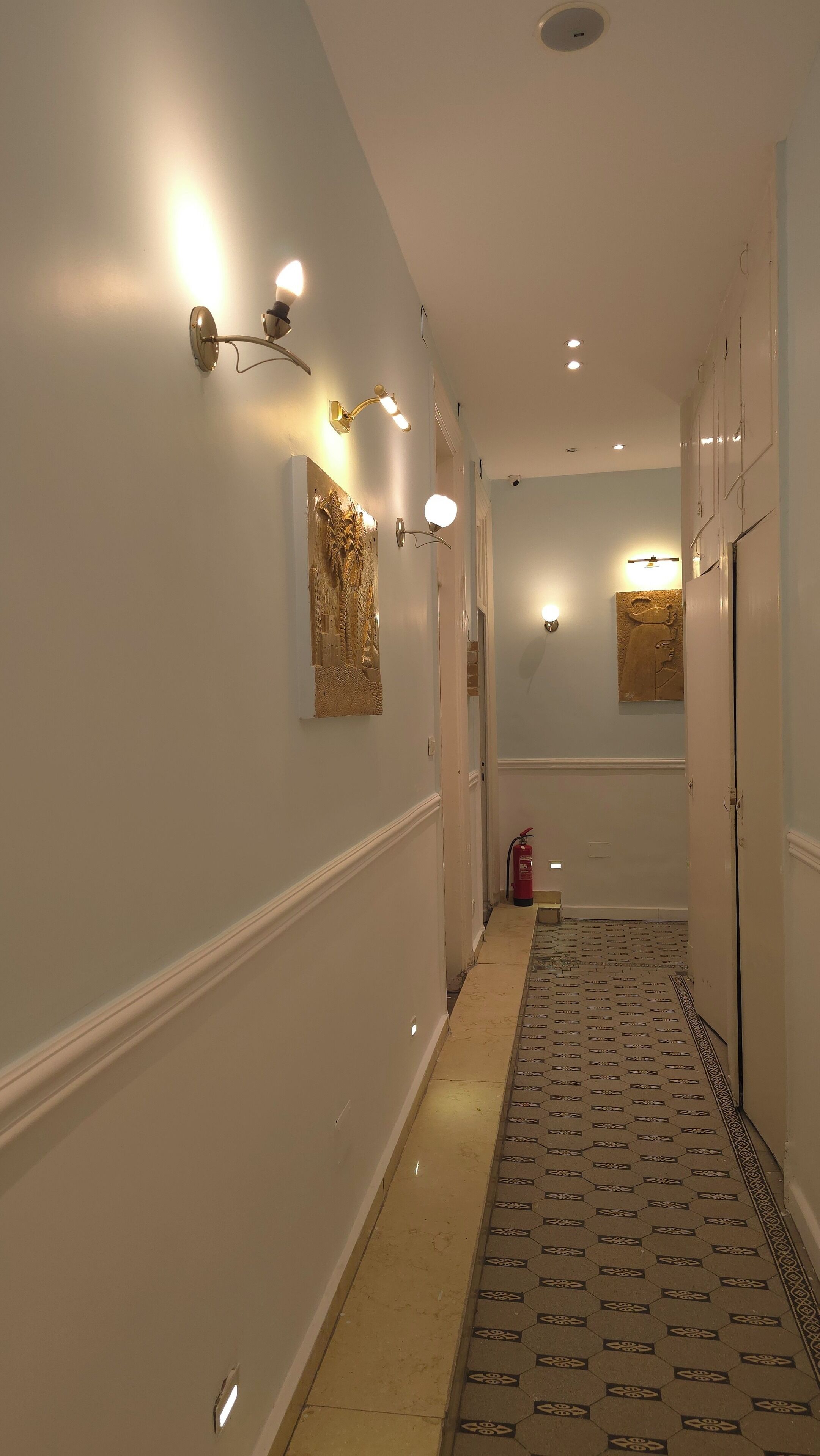 A hallway with white walls, decorative wall sconces, a patterned floor, and a fire extinguisher.