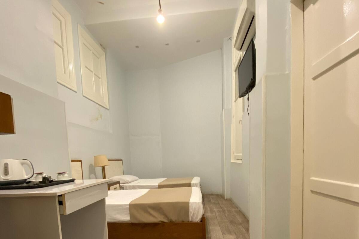 Un dormitorio con cama, mesita de noche y escritorio con silla. Se observa una puerta y una ventana en la pared.