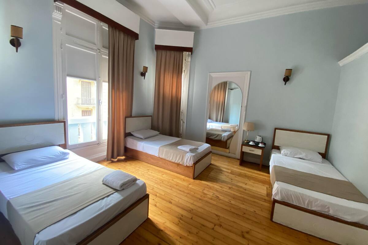 Habitación de hotel con tres camas, suelo de madera y un ventanal grande con cortinas.