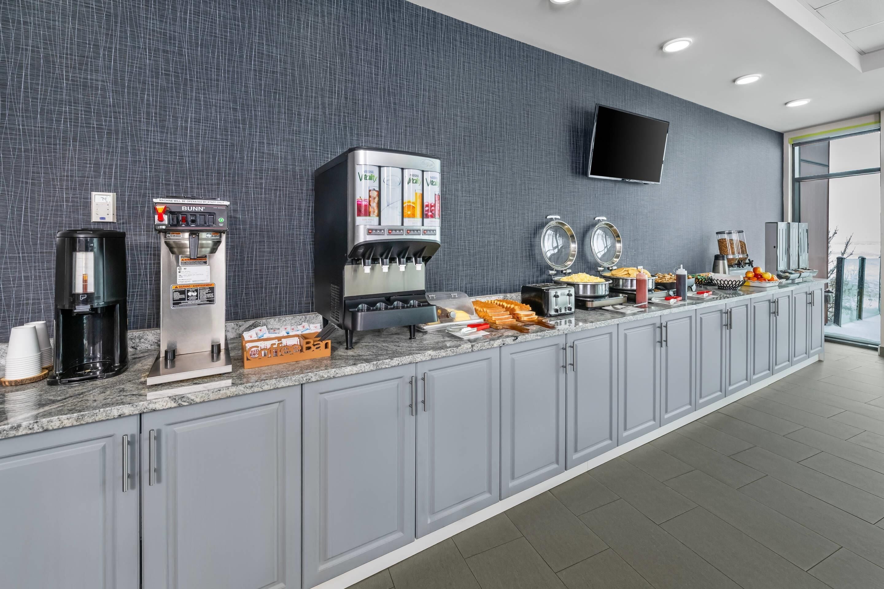 Un espace cuisine moderne équipé d’une machine à café, d’un distributeur de jus et d’un bar pour le petit-déjeuner, où sont proposés divers aliments.
