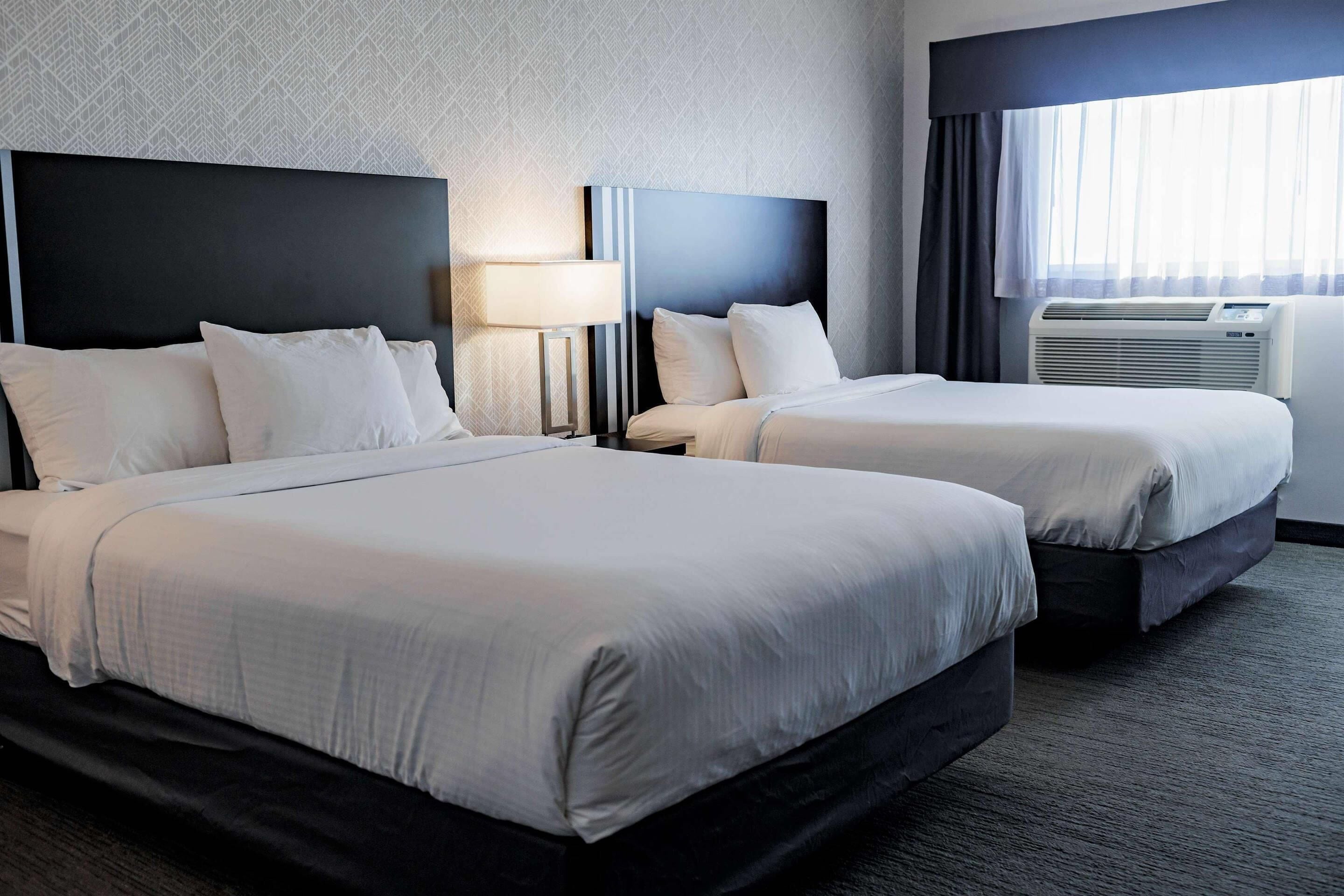 A hotel room with two beds, a nightstand with a lamp, an air conditioning unit, and a window with curtains.