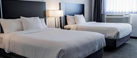 A hotel room with two beds, a nightstand with a lamp, an air conditioning unit, and a window with curtains.