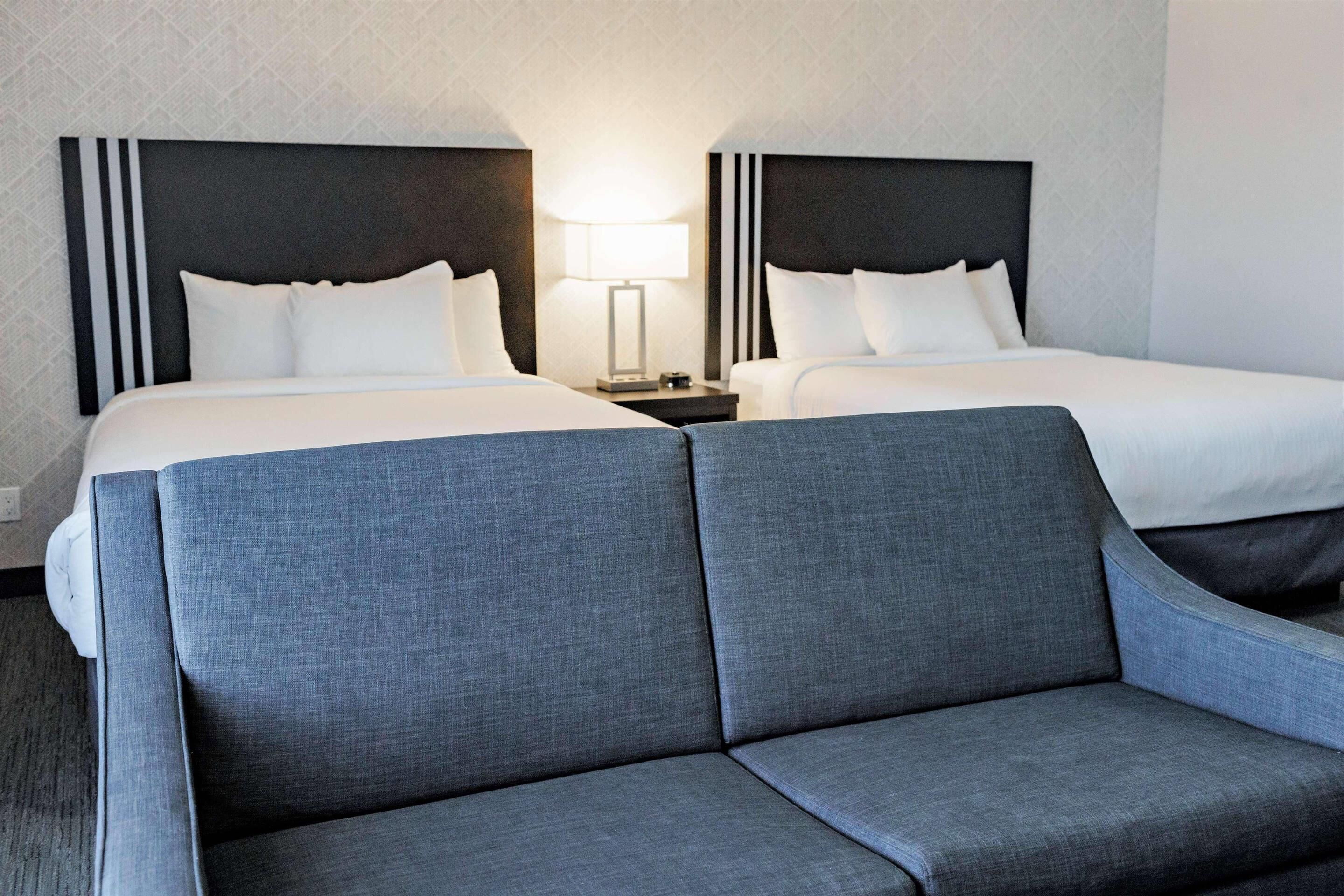 Una habitación de hotel con dos camas, una mesita de noche con lámpara y un sofá gris.