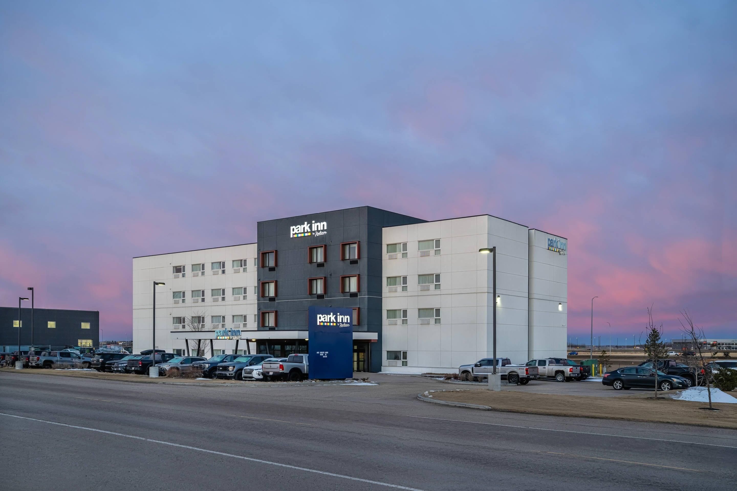 Un hotel de varios pisos con el cartel 'Park Inn', un estacionamiento y cielo despejado.