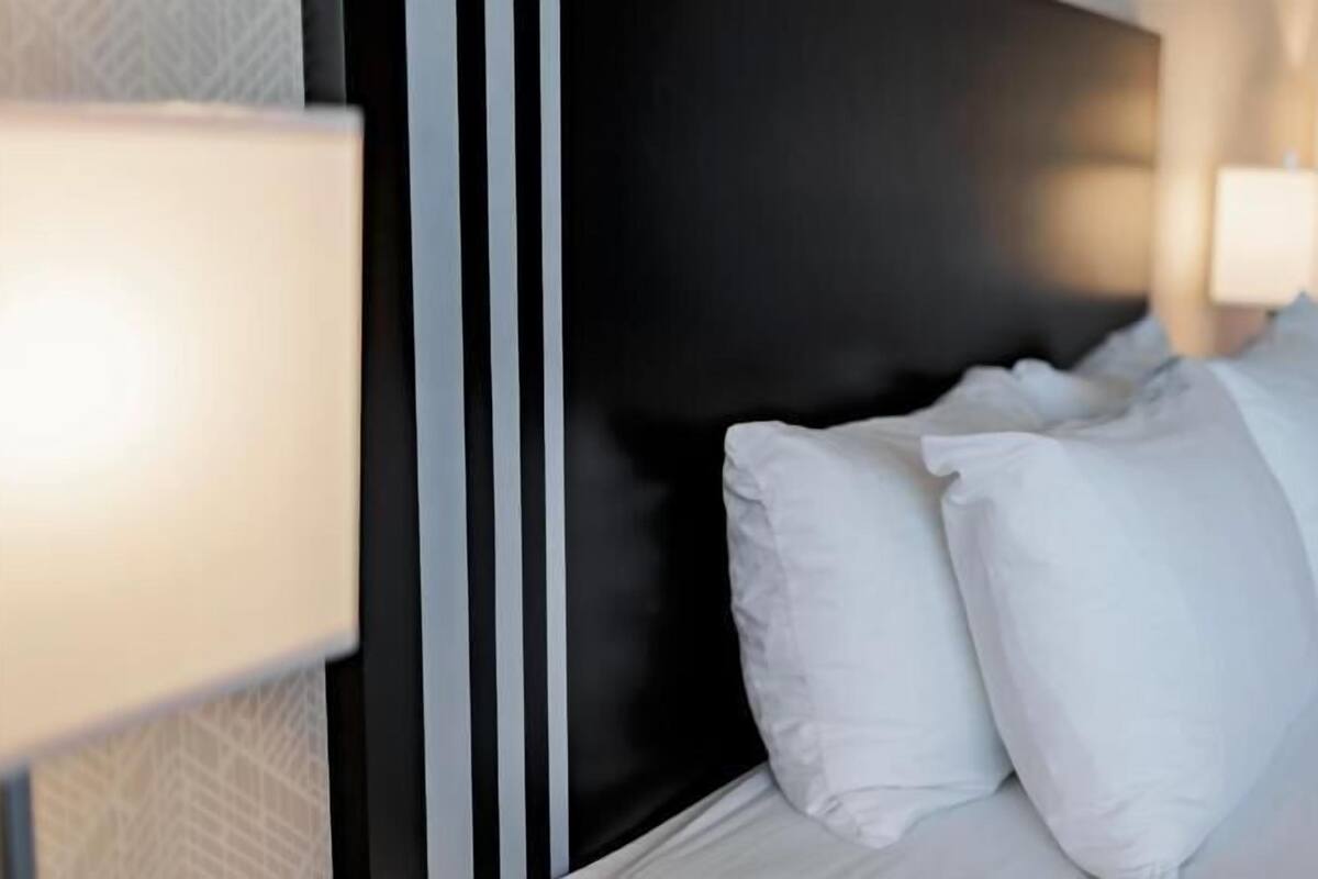 A hotel room with a bed, white pillows, a dark headboard, and two wall-mounted lamps.