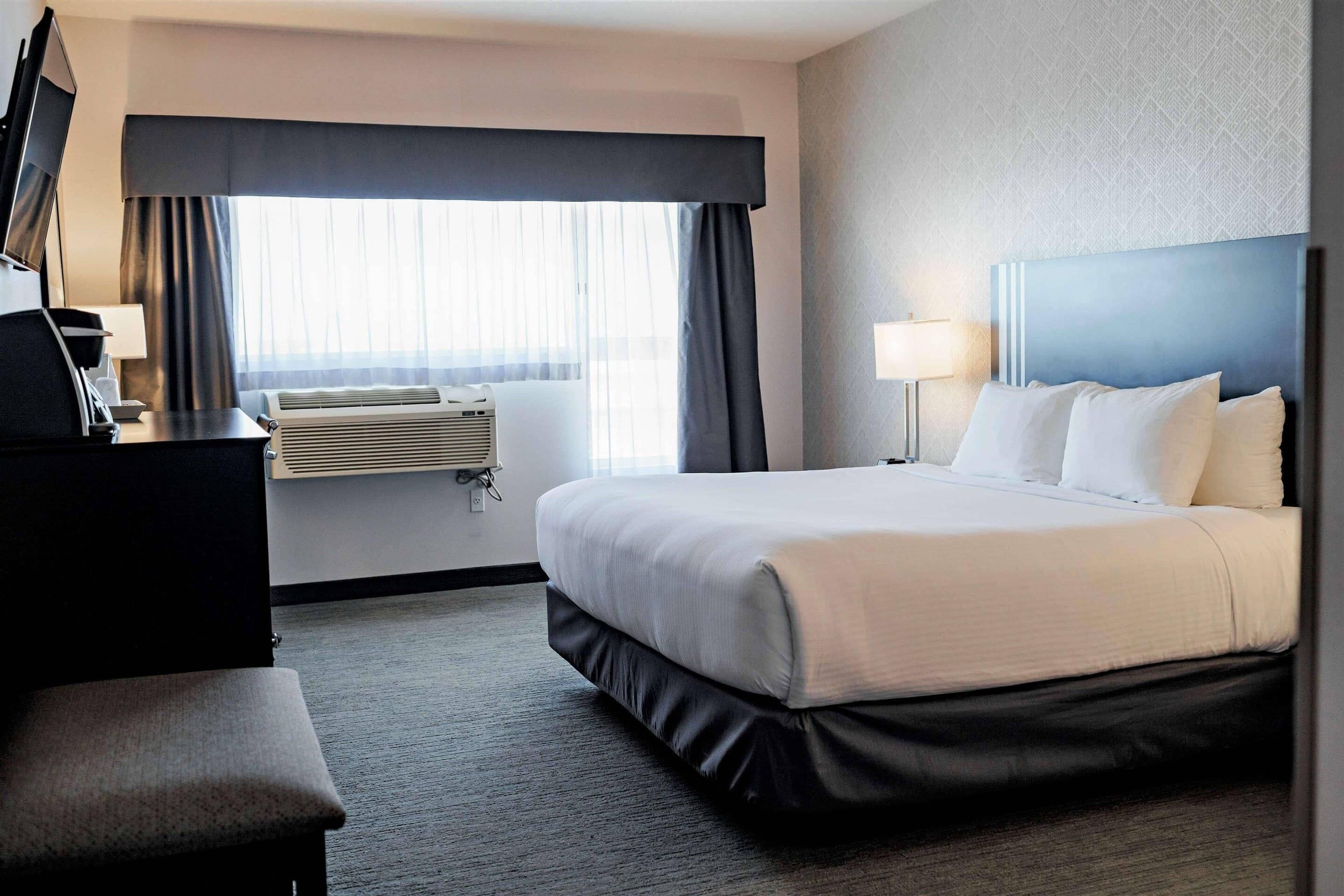 A hotel room with a bed, a desk, a chair, a lamp, and a window with curtains.