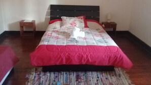 Una cama bien hecha con ropa de cama floral, un detalle rojo y una toalla blanca. Hay dos mesitas de noche, una con una lámpara.