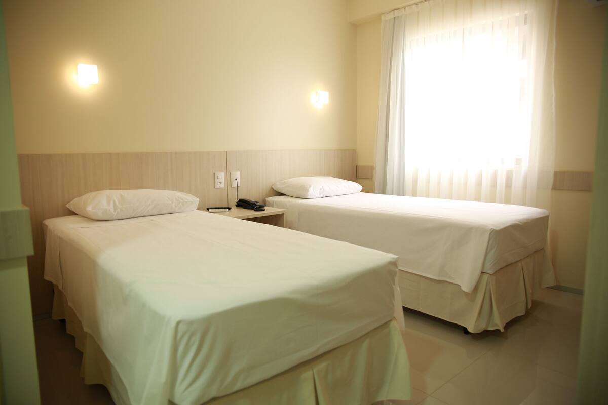 Habitación de hotel con dos camas individuales, una mesita de noche con teléfono y una ventana con cortinas.