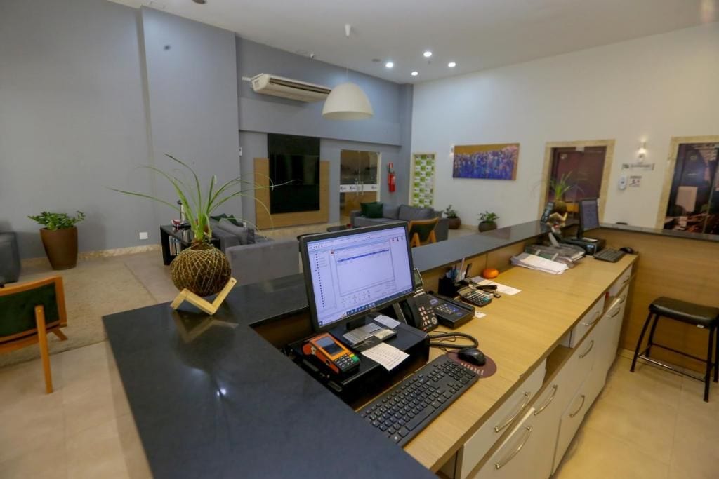 A modern reception desk with a computer, telephone, and various office supplies. There are plants, a couch, and a painting on the wall.