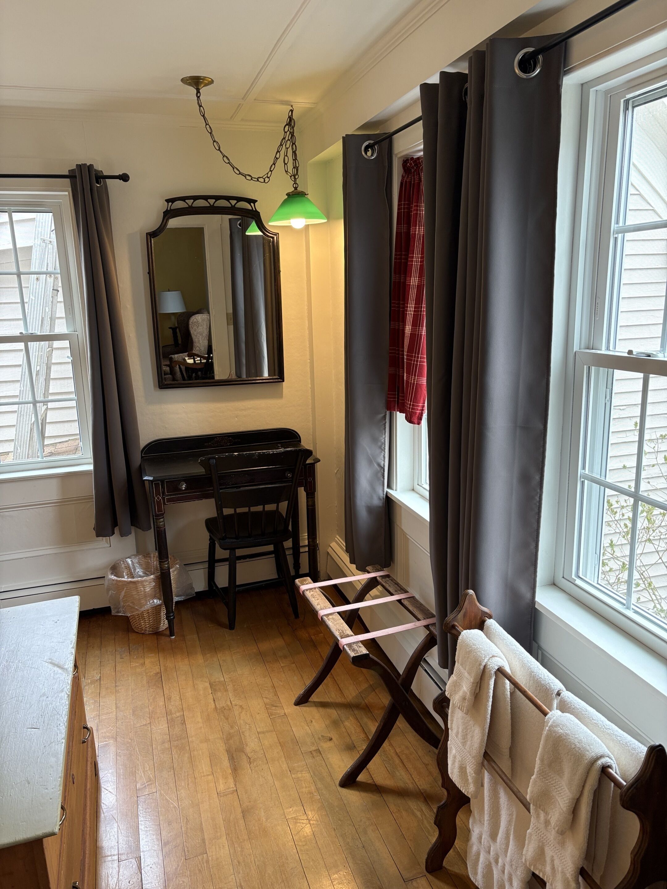 A room with a wooden desk, a chair, a mirror, a hanging light fixture, a window with curtains, and a towel rack.