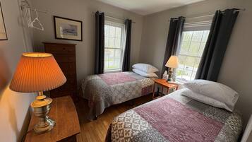 A room with two beds, a wooden dresser, a lamp, a framed picture, and a window with curtains.