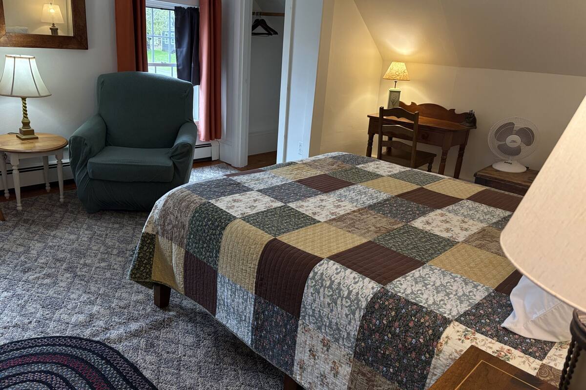Ein Schlafzimmer mit Bett, Stuhl, Lampe, Ventilator und einem Fenster mit Vorhängen.