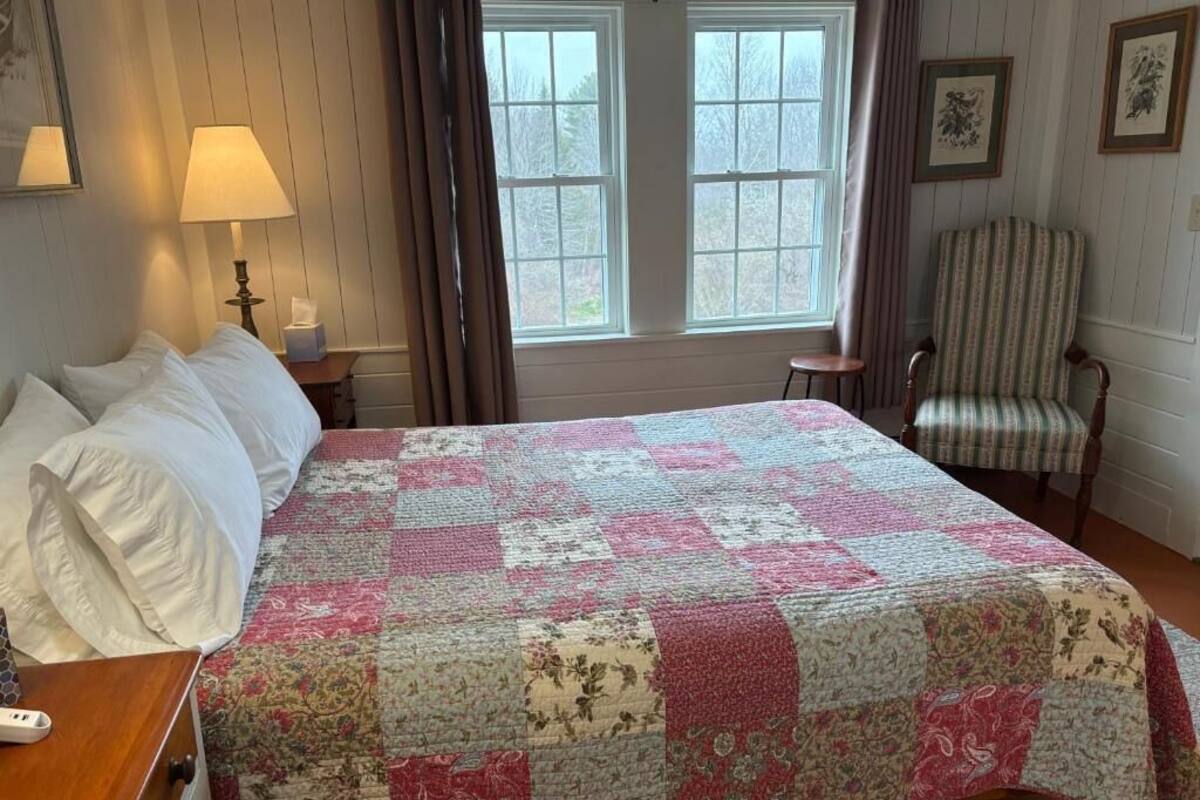 Ein Schlafzimmer mit einem großen Bett, einem Sessel, einem Nachttisch, einer Lampe und zwei Fenstern.