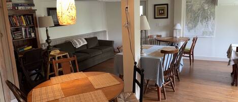 A dining area with a round wooden table, chairs, and placemats. There is a sofa and a bookshelf in the background.