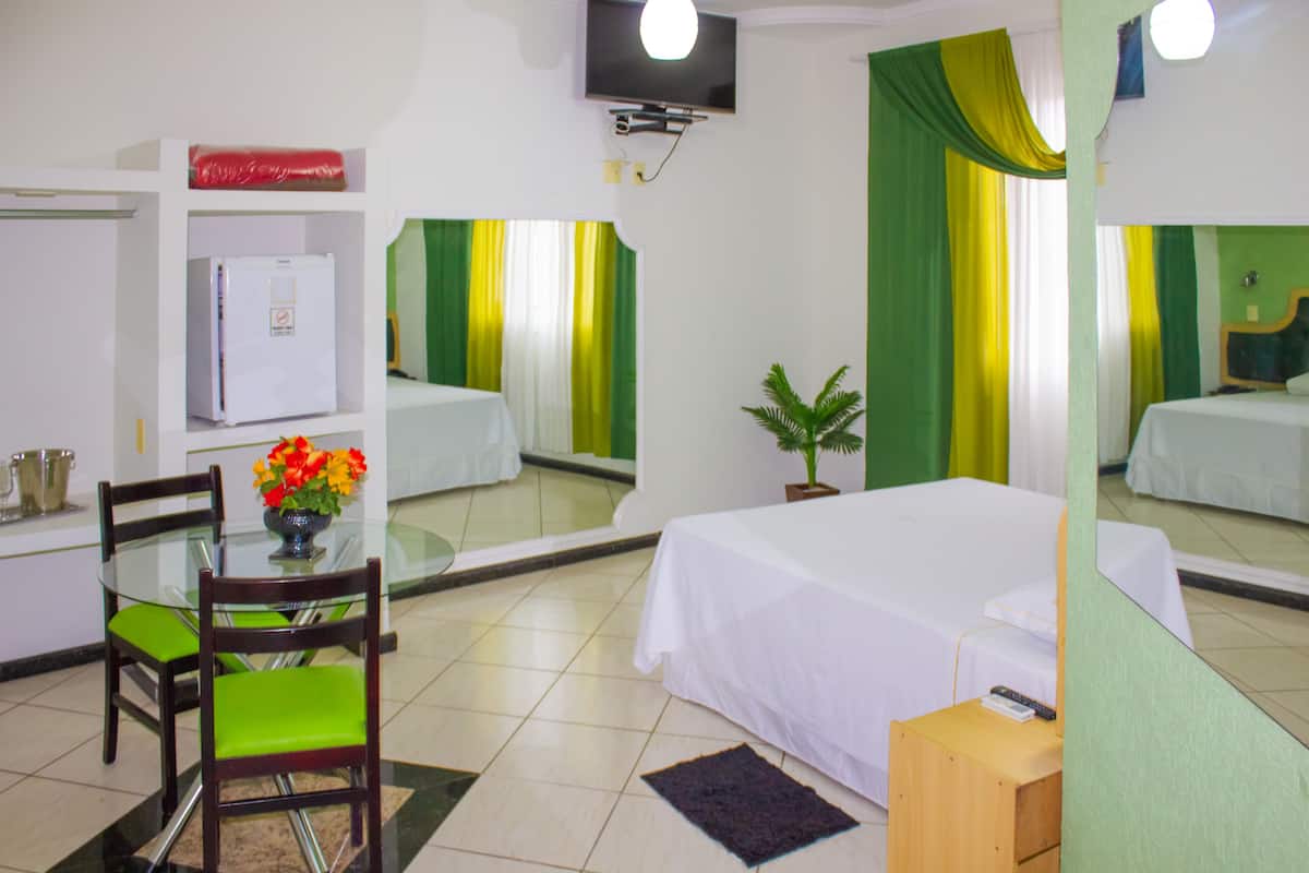 Quarto de hotel com cama, escrivaninha com cadeira, pequena mesa com um vaso de flores, televisão e frigorífico.