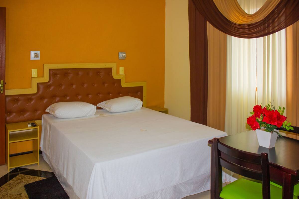 Quarto de hotel com uma cama grande, uma mesinha de cabeceira, uma cadeira, uma mesinha pequena com um vaso de flores e cortinas.