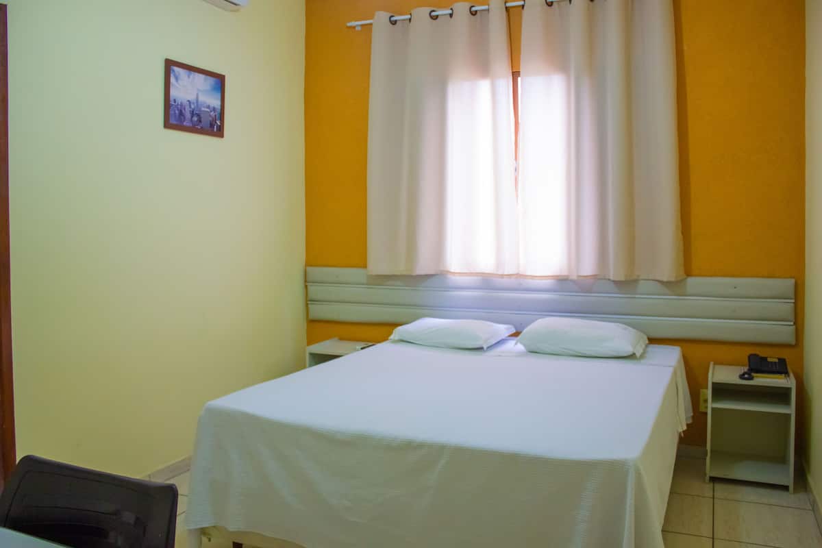 Uma cama de solteiro com um cobre-leito branco e duas almofadas, um criado-mudo com telefone e um quadro na parede.