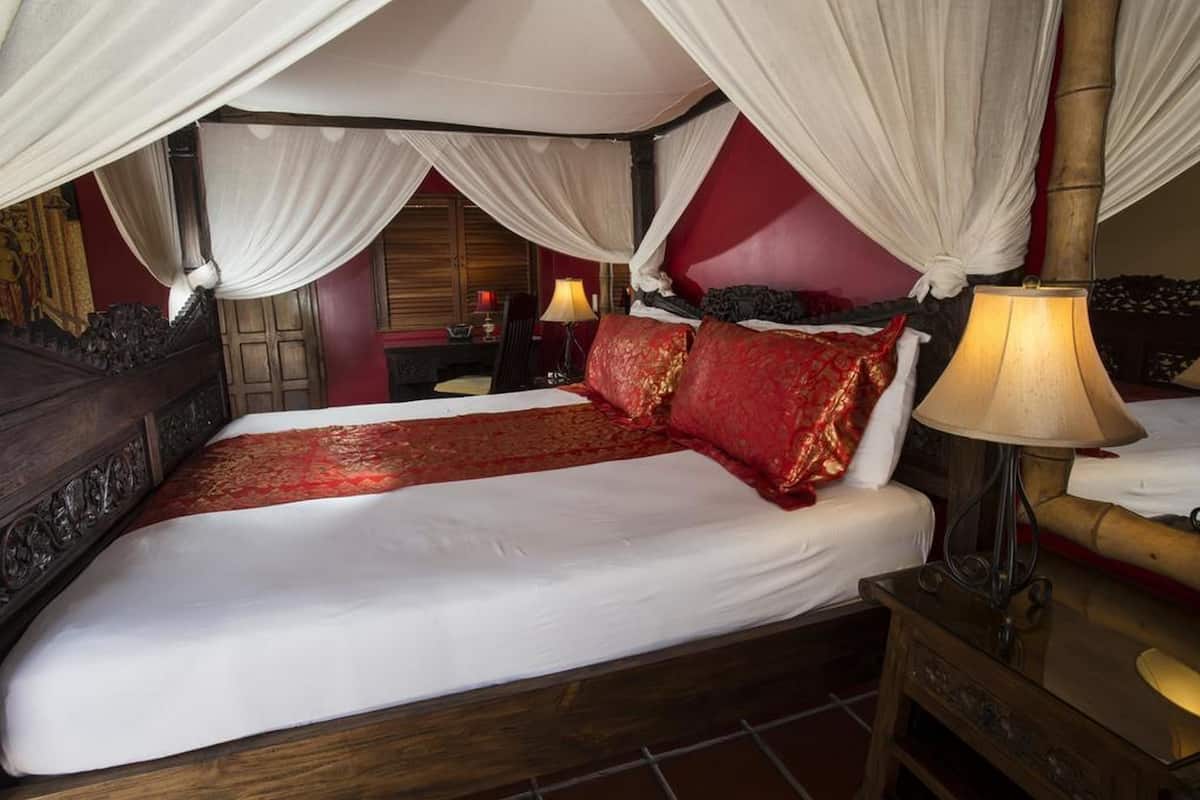 Uma cama de dossel com roupa de cama branca e detalhes vermelhos, uma mesa de cabeceira com abajur e um criado-mudo de madeira.