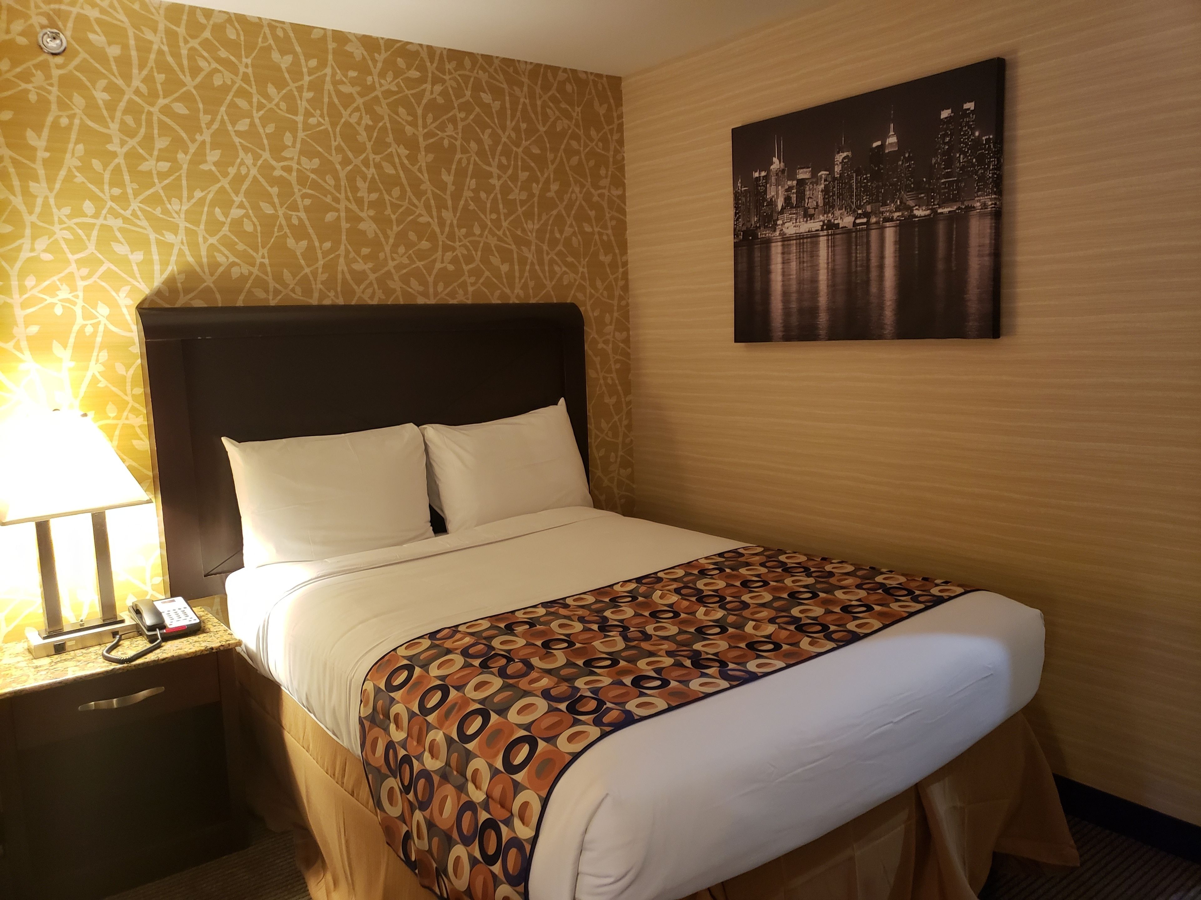 Een hotelkamer met een bed, een nachtkastje met een lamp, een telefoon en een stadslandschap schilderij aan de muur.