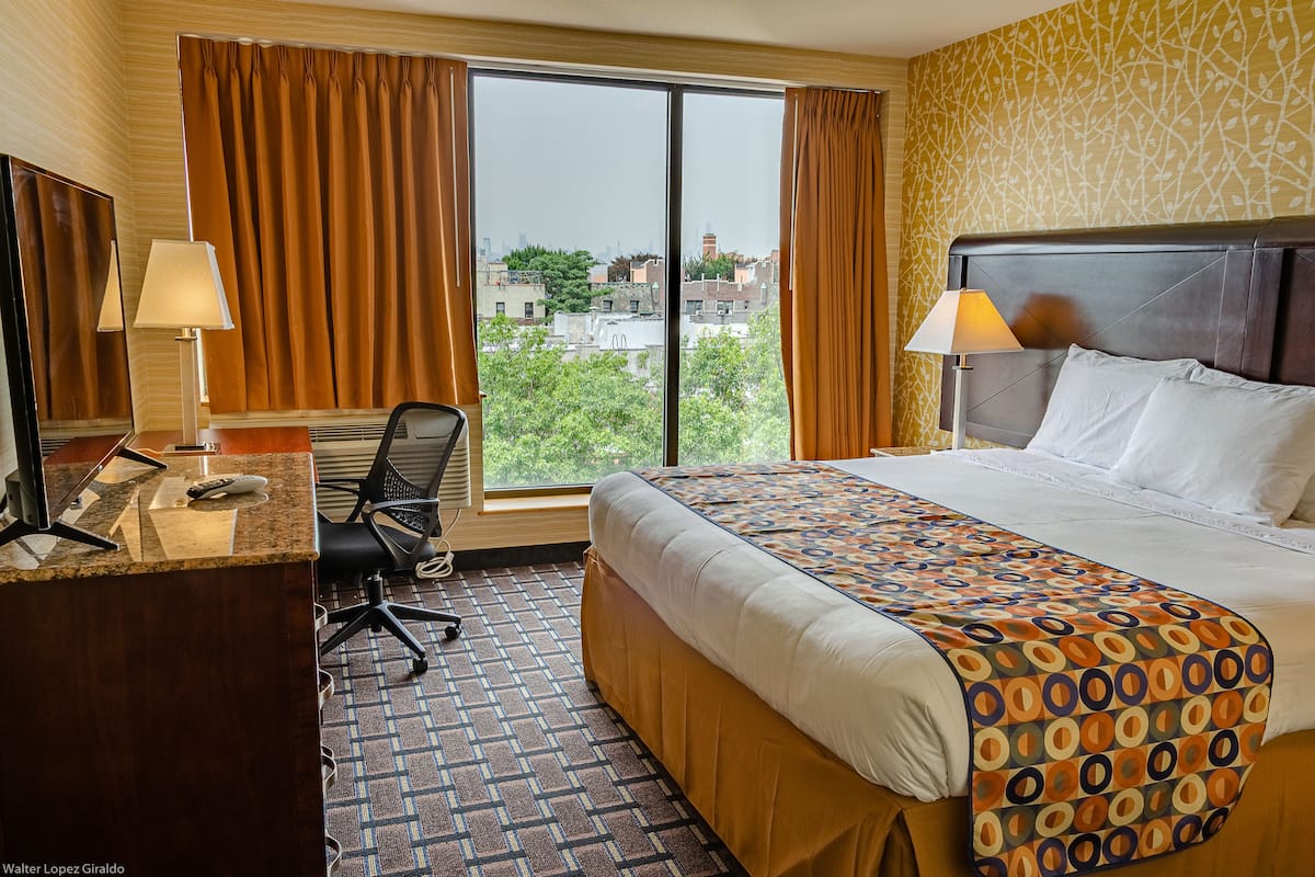 A hotel room with a large bed, a desk with a chair, a window with curtains, and a city view.