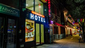 Un hotel con un letrero de neón, una farmacia al otro lado de la calle y una dirección visible.
