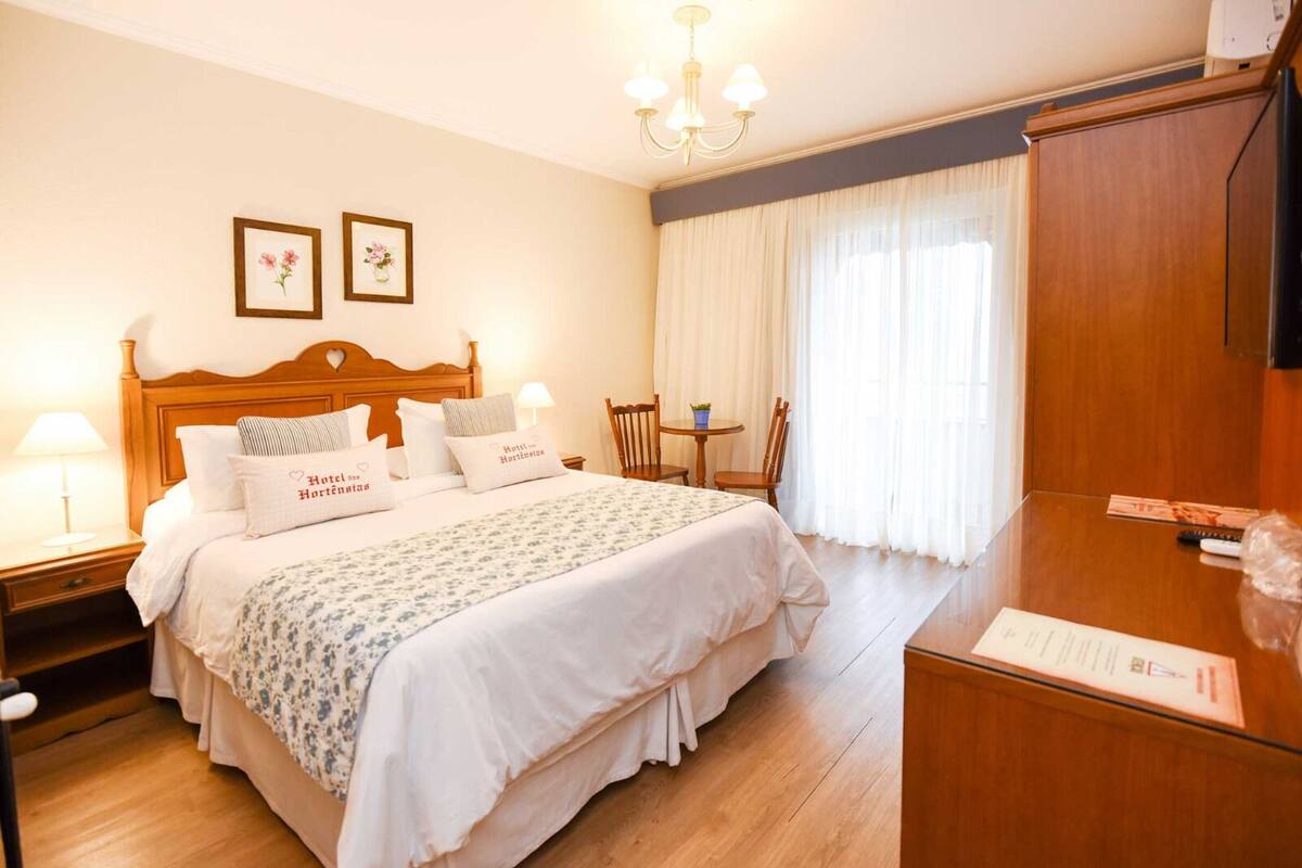 Habitación de hotel con una cama grande, cabecera de madera, dos mesitas de noche con lámparas, armario de madera y una pequeña mesa con sillas.