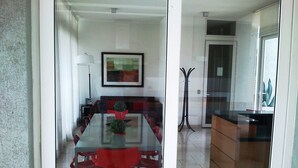 Un comedor moderno con una mesa de vidrio, sillas rojas, una obra enmarcada en la pared y una cocina visible a través del vidrio.