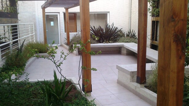 Un espacio al aire libre techado, con suelo de hormigón, pilares de madera y diversas plantas.