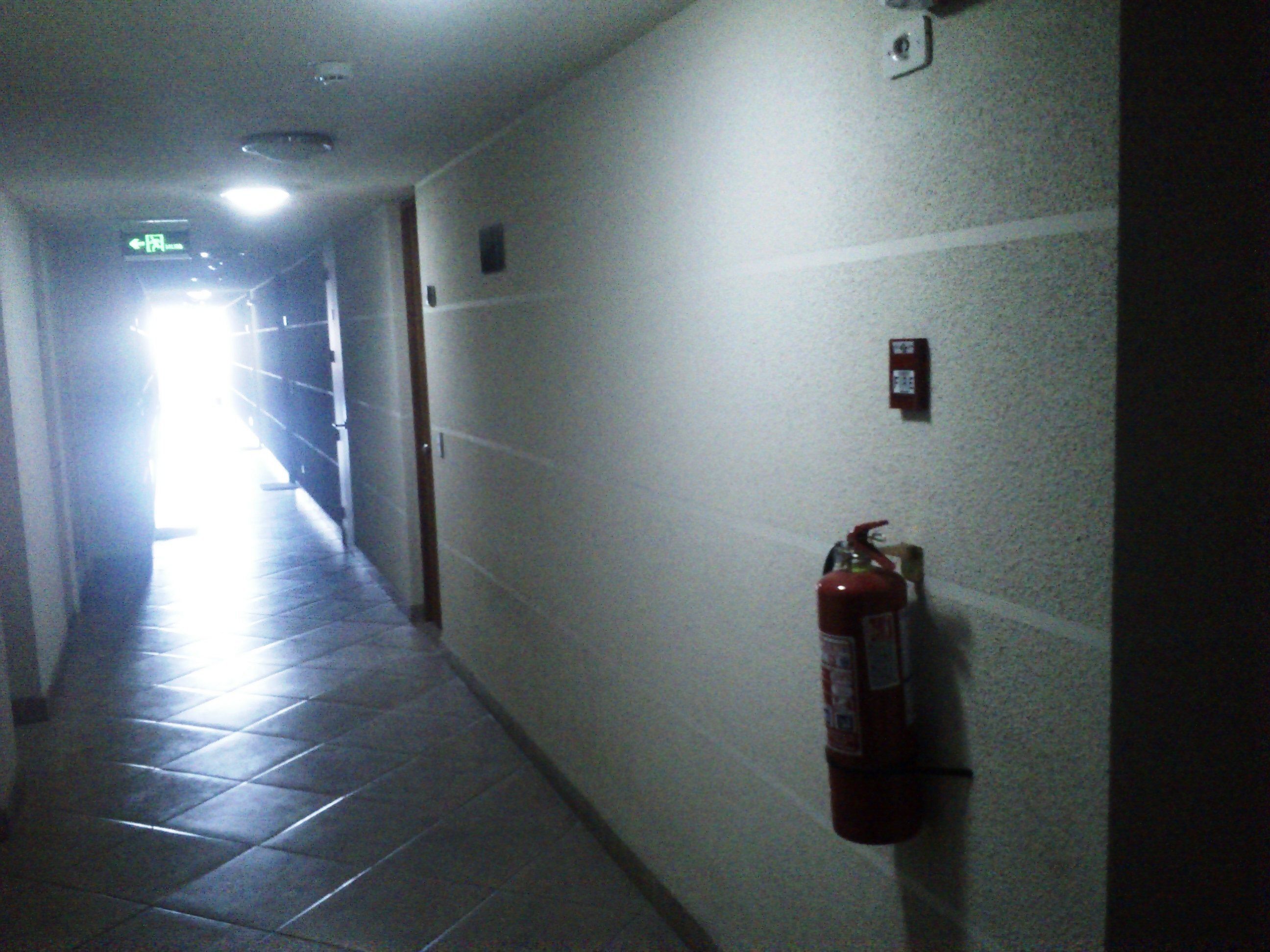 A hallway with a fire extinguisher mounted on the wall, a light fixture on the ceiling, and a door at the end.