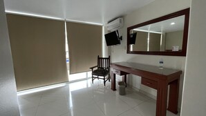 Habitación de hotel con escritorio de madera, una silla, un espejo grande, televisión y ventana con persianas.