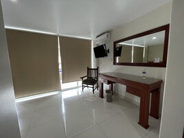 A hotel room with a wooden desk, a chair, a large mirror, a television, and a window with blinds.