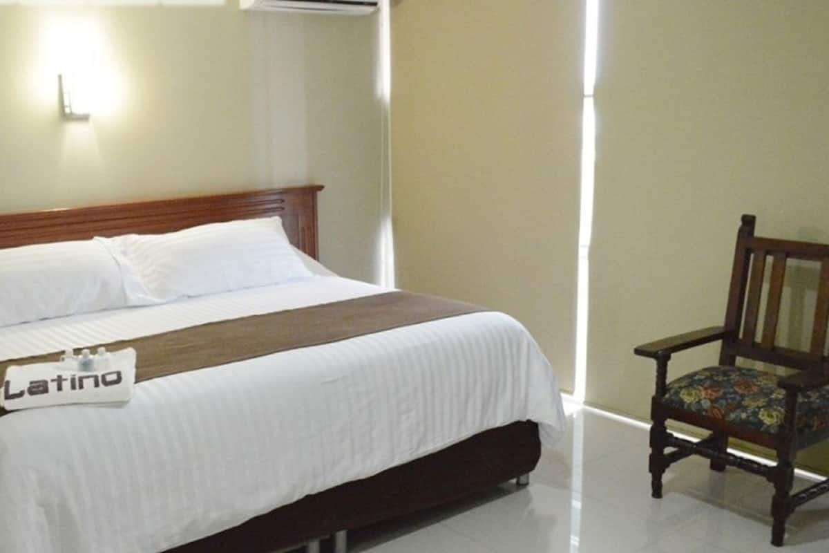 A neatly made bed with a white comforter and a brown blanket, a wooden nightstand with a lamp, and a wooden chair with a floral cushion.