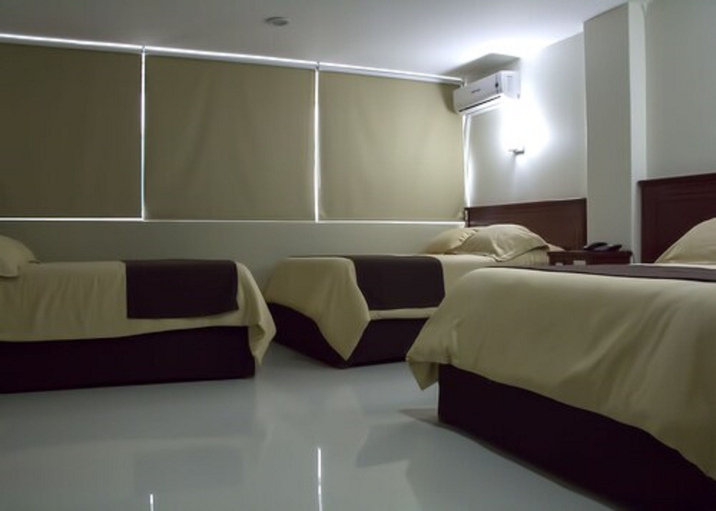 Habitación de hotel con tres camas alineadas, un ventanal con persianas y un equipo de aire acondicionado instalado en la pared.
