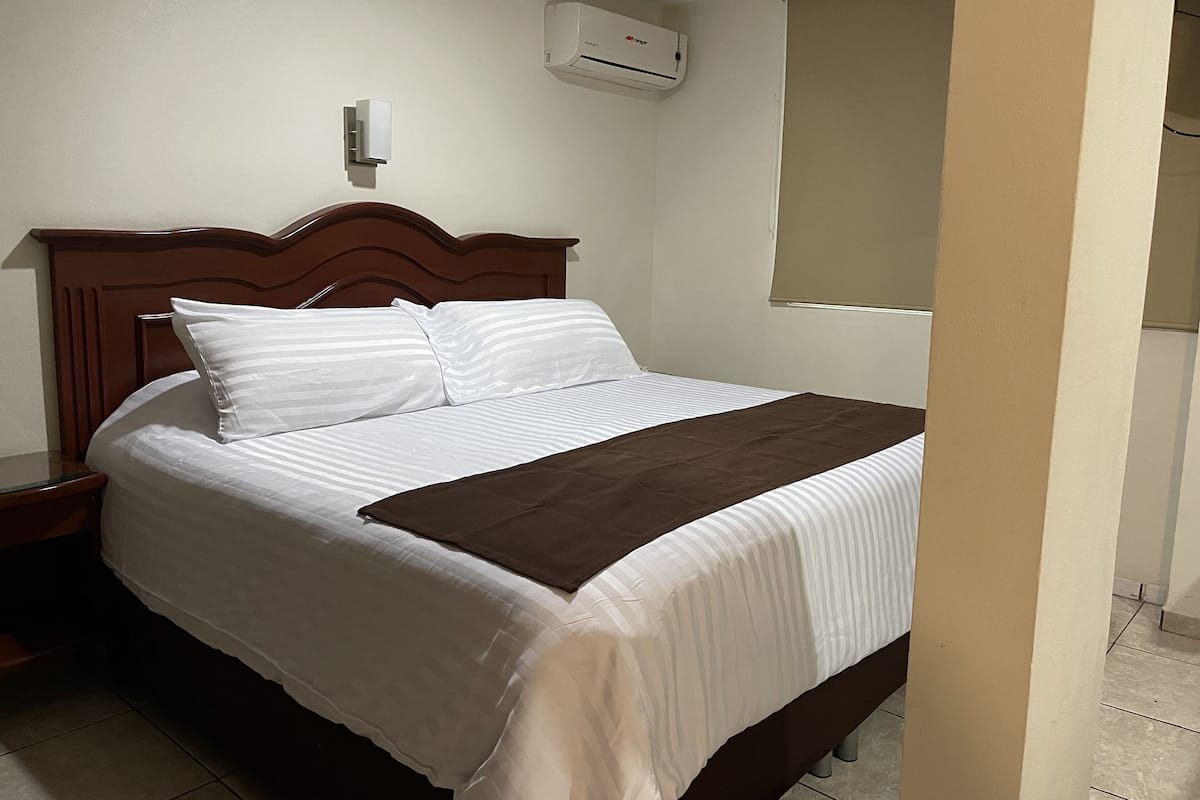 A neatly made bed with white and brown linens, a wooden headboard, and a wall-mounted air conditioner.