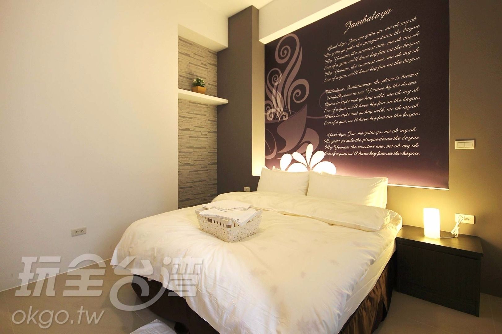 A modern bedroom with a large bed, a nightstand, a wall-mounted shelf, and a decorative wall piece.