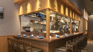 A restaurant with a long bar, wooden chairs, and a kitchen area visible through the bar.