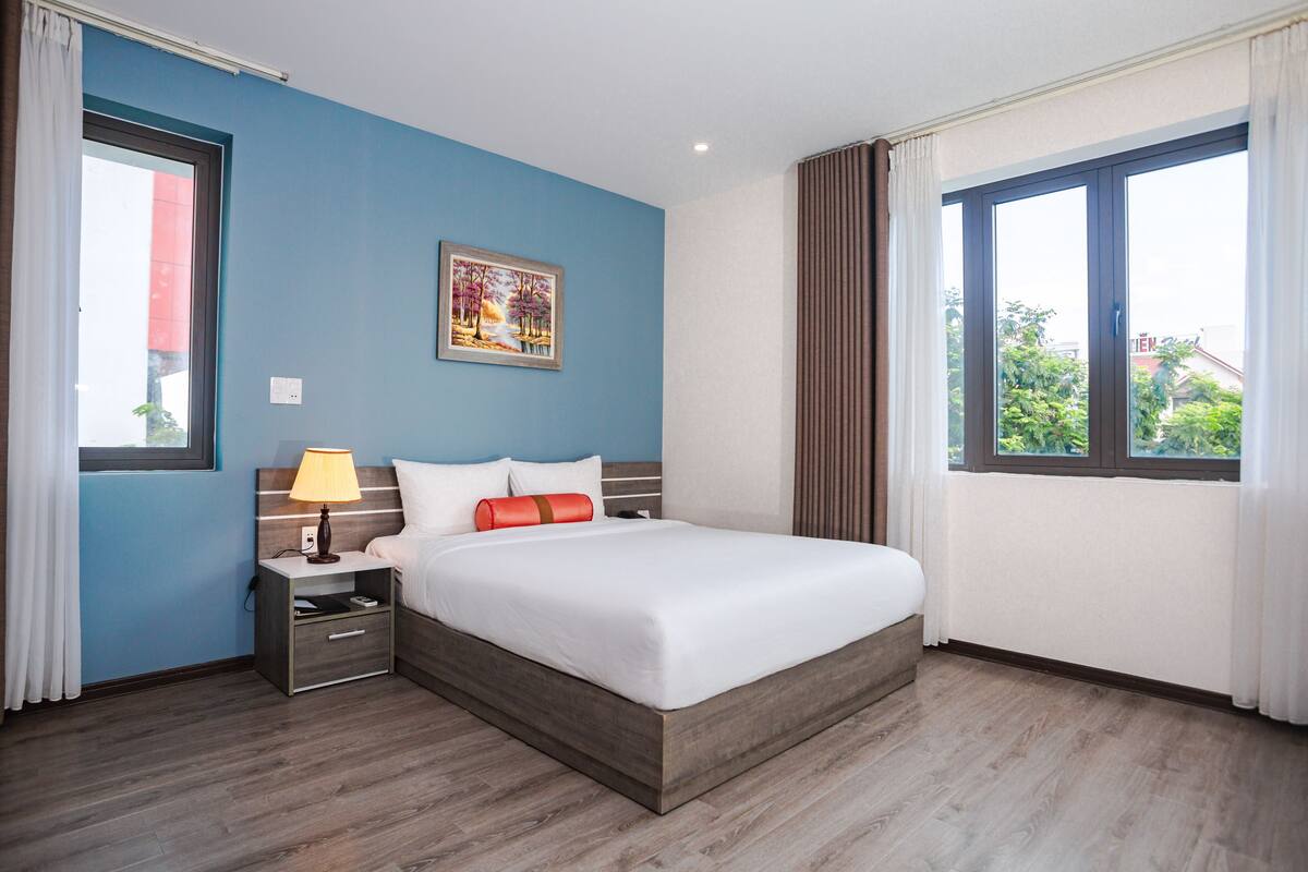 Ein Hotelzimmer mit Bett, Nachttisch, Lampe, einem Bild an der Wand und einem Fenster mit Vorhängen.