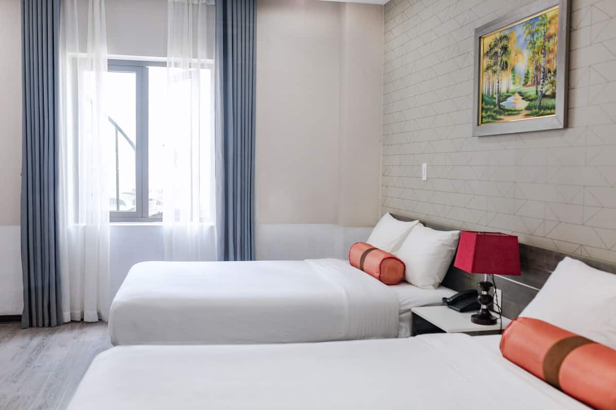 Ein Hotelzimmer mit zwei Betten, einem Fenster mit durchsichtigen Vorhängen, einem Gemälde an der Wand und einem Nachttisch mit Lampe und Telefon.