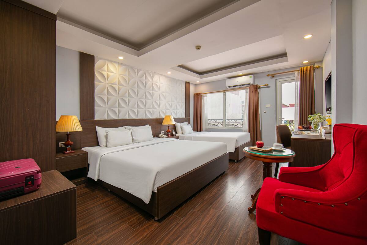 Hotelkamer met een groot bed, twee nachtkastjes, een rode fauteuil, een klein rond tafeltje met een vaas, en een raam met gordijnen.
