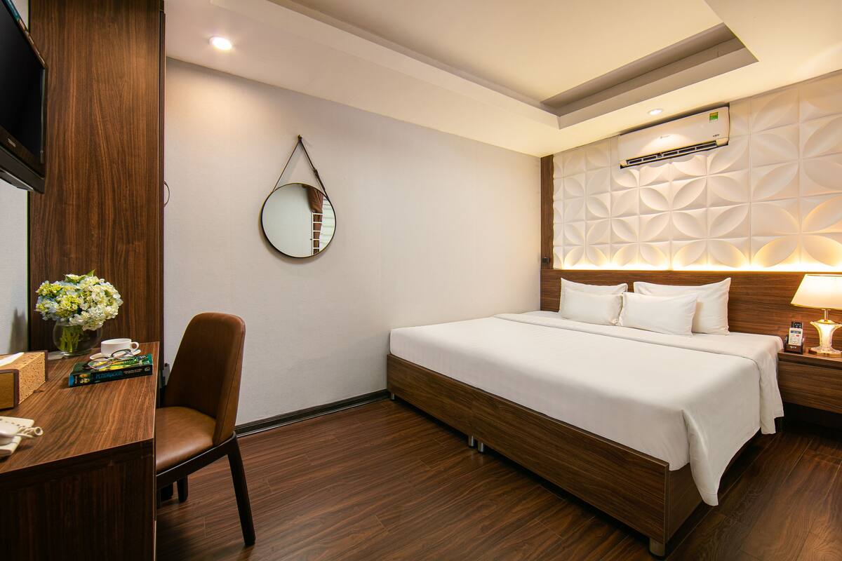 Een hotelkamer met een houten bed, een bureau met stoel, een ronde spiegel en een wandairconditioner.