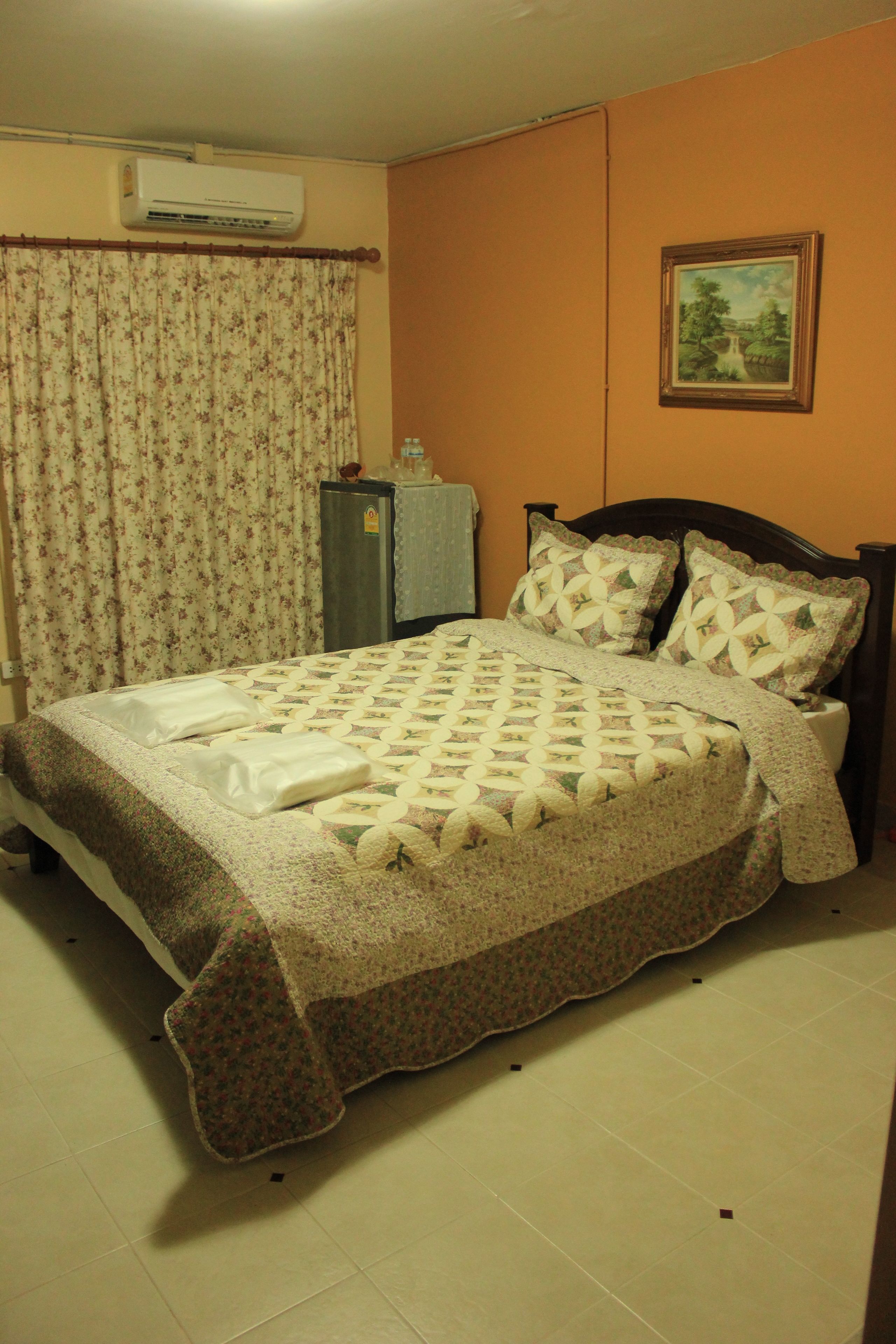 A bedroom with a bed, floral curtains, a painting on the wall, and an air conditioning unit.